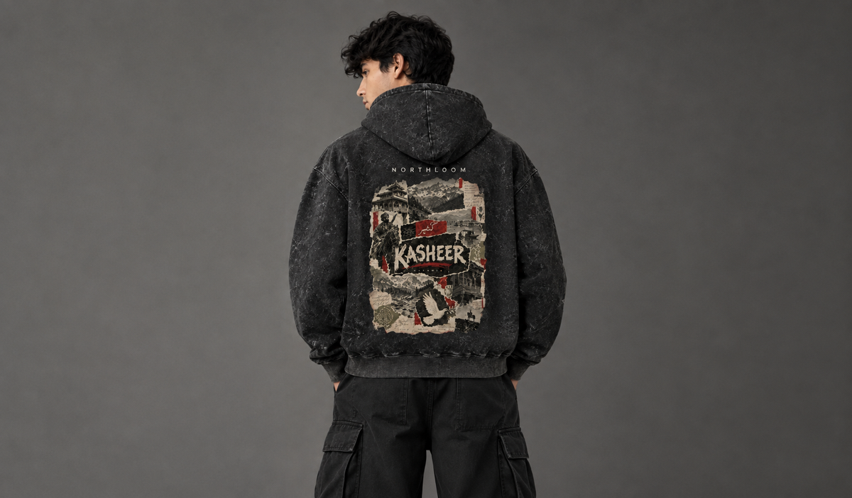 A person shown from the back wearing a dark washed hoodie with a large graphic print on the back reading 'KASHEER', paired with black cargo pants against a dark gray studio background.